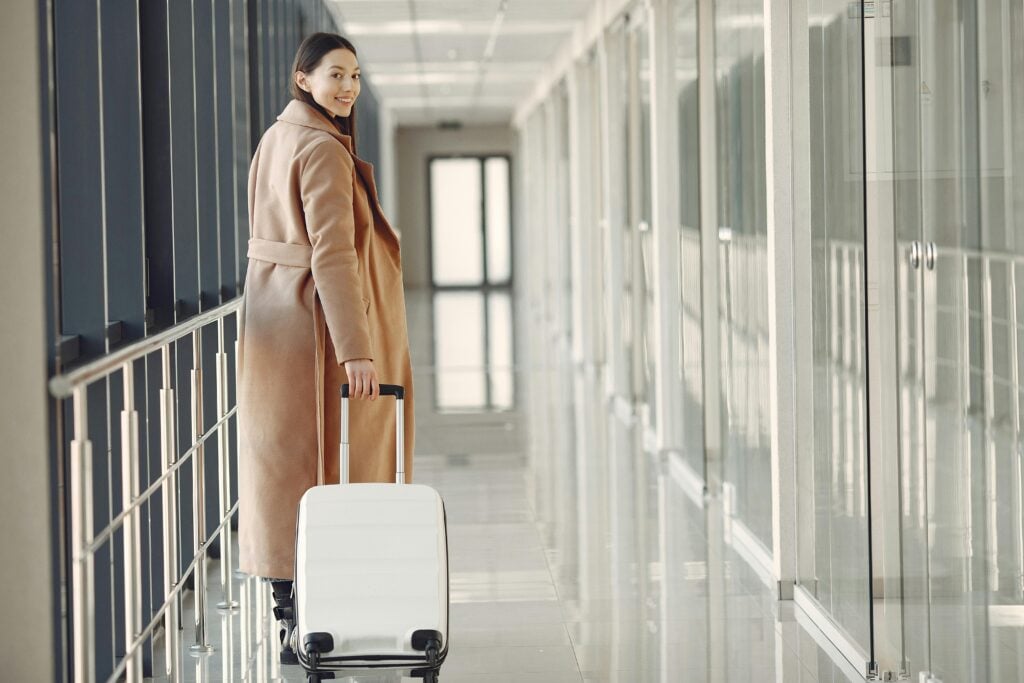 Stylish happy traveler with suitcase in airport hallway